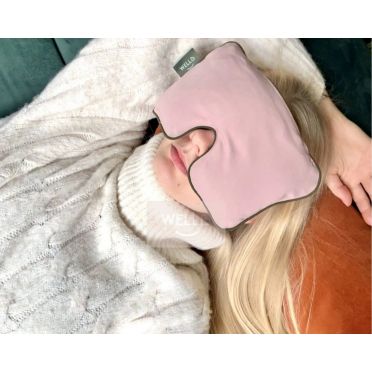 Kobieta w białym swetrze odpoczywa na kanapie z różową poduszką na oczy, głowa oparta na ciemnej poduszce, tło rozmyte.